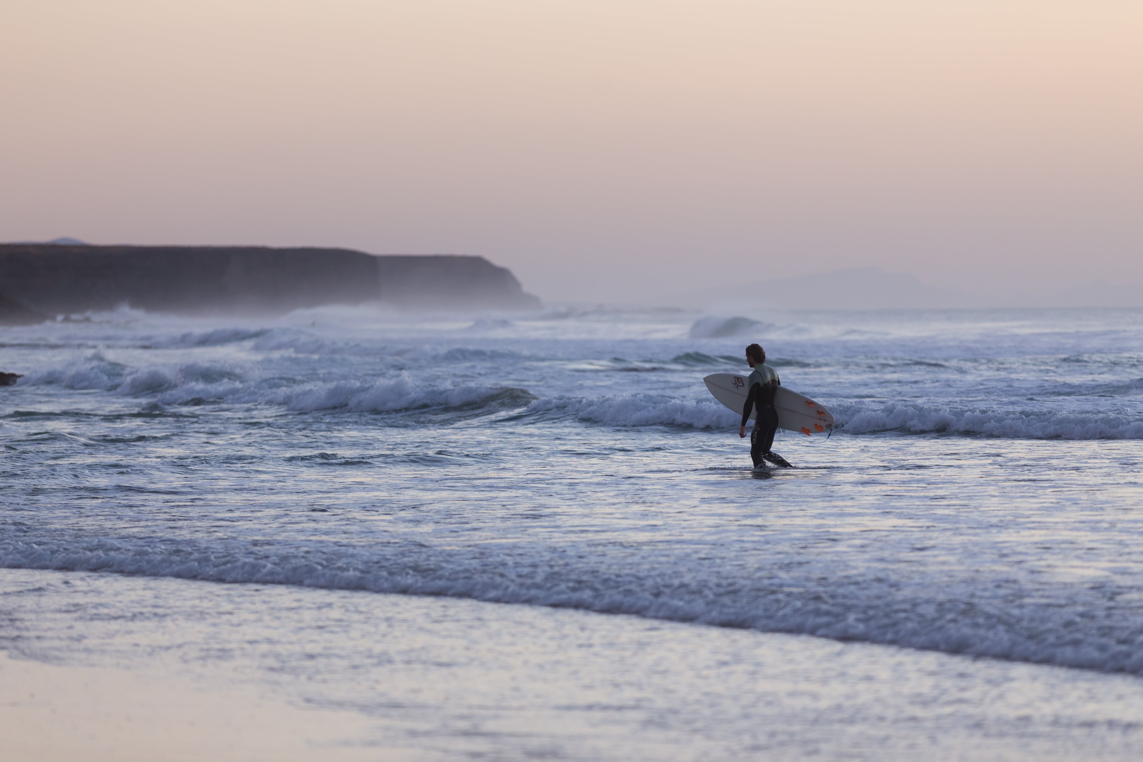 Surfeur entrant dans l'eau à El Cotillo au coucher du soleil, avec de douces vagues et des tons violets dans le ciel, reflétant la tranquillité du moment.